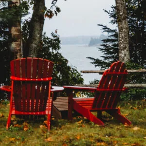 Two red Adirondack chairs facing a misty lake, framed by evergreen trees and a rustic wooden railing.